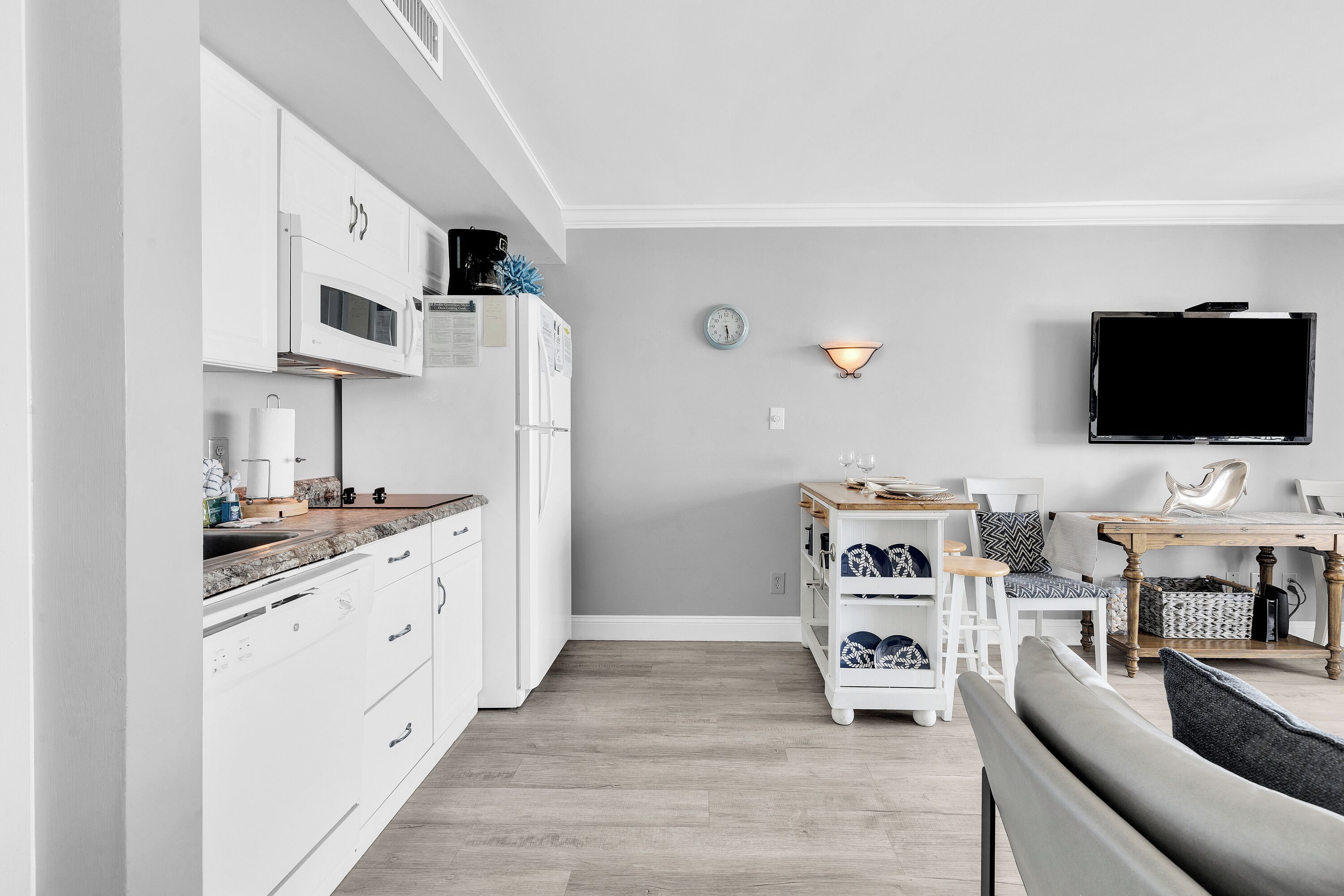 Una cocina moderna con armarios blancos, un microondas y una cafetera. Hay un comedor con una mesa y sillas, y un televisor montado en la pared.