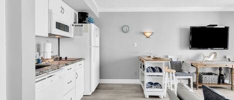 Una cocina moderna con armarios blancos, un microondas y una cafetera. Hay un comedor con una mesa y sillas, y un televisor montado en la pared.