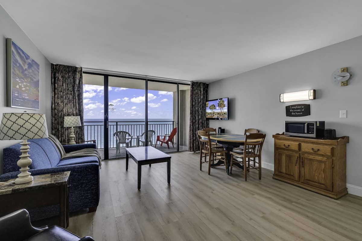A living room with a sofa, a dining table with chairs, a TV, a wooden cabinet, and a view of the ocean through large windows.