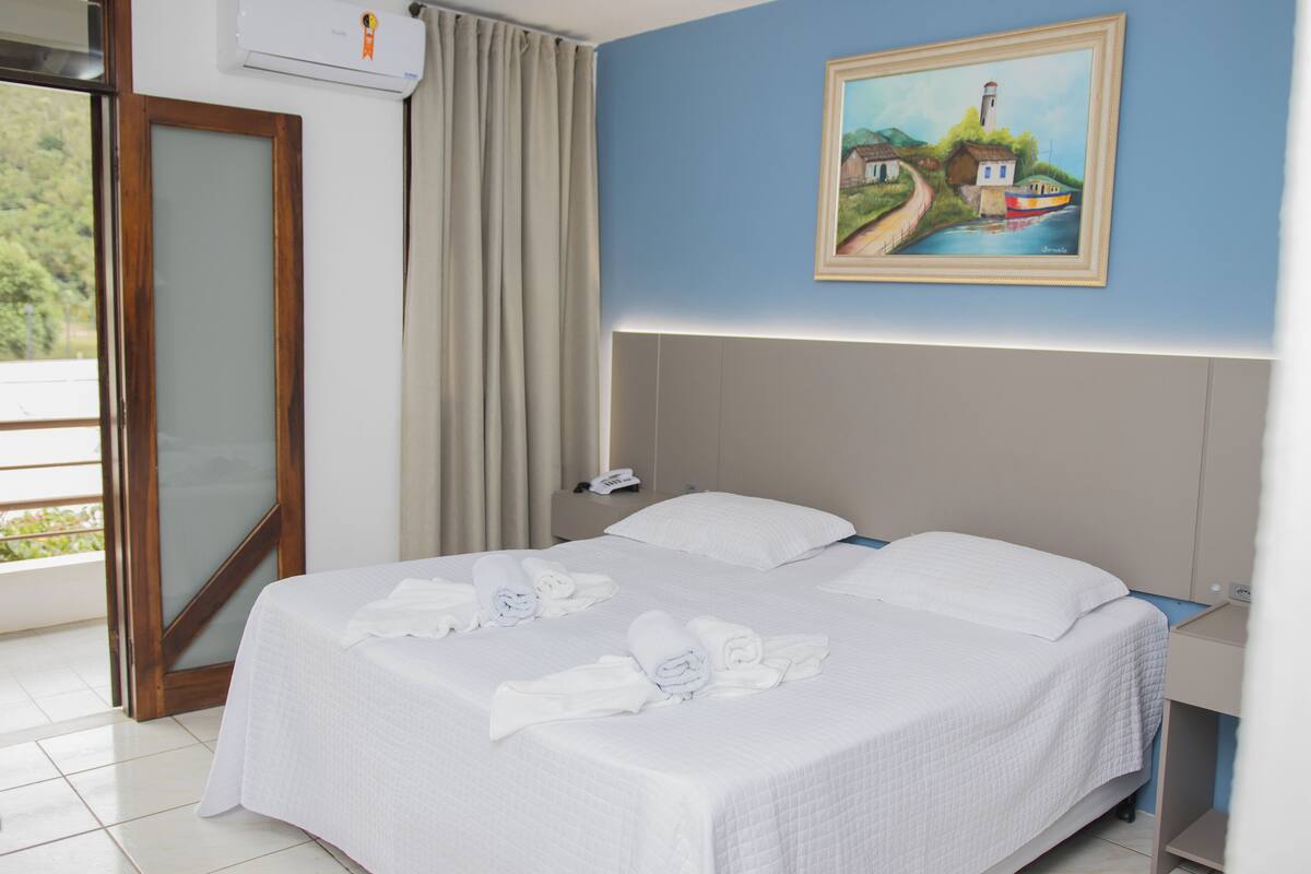 A hotel room with a bed, a painting on the wall, a nightstand, and a view of the outdoors.