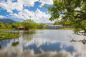 A serene lake with a reflection of the sky and surrounding greenery, a building with a red roof, and mountains in the background. - Hotel Termas do Lago (Gravatal)
