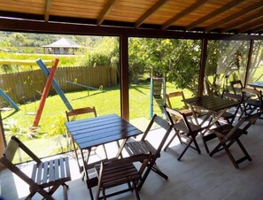 An outdoor patio with wooden tables and chairs, a thatched-roof structure, and a view of a grassy area with a fence and trees. - Pousada A Cabana (Garopaba)