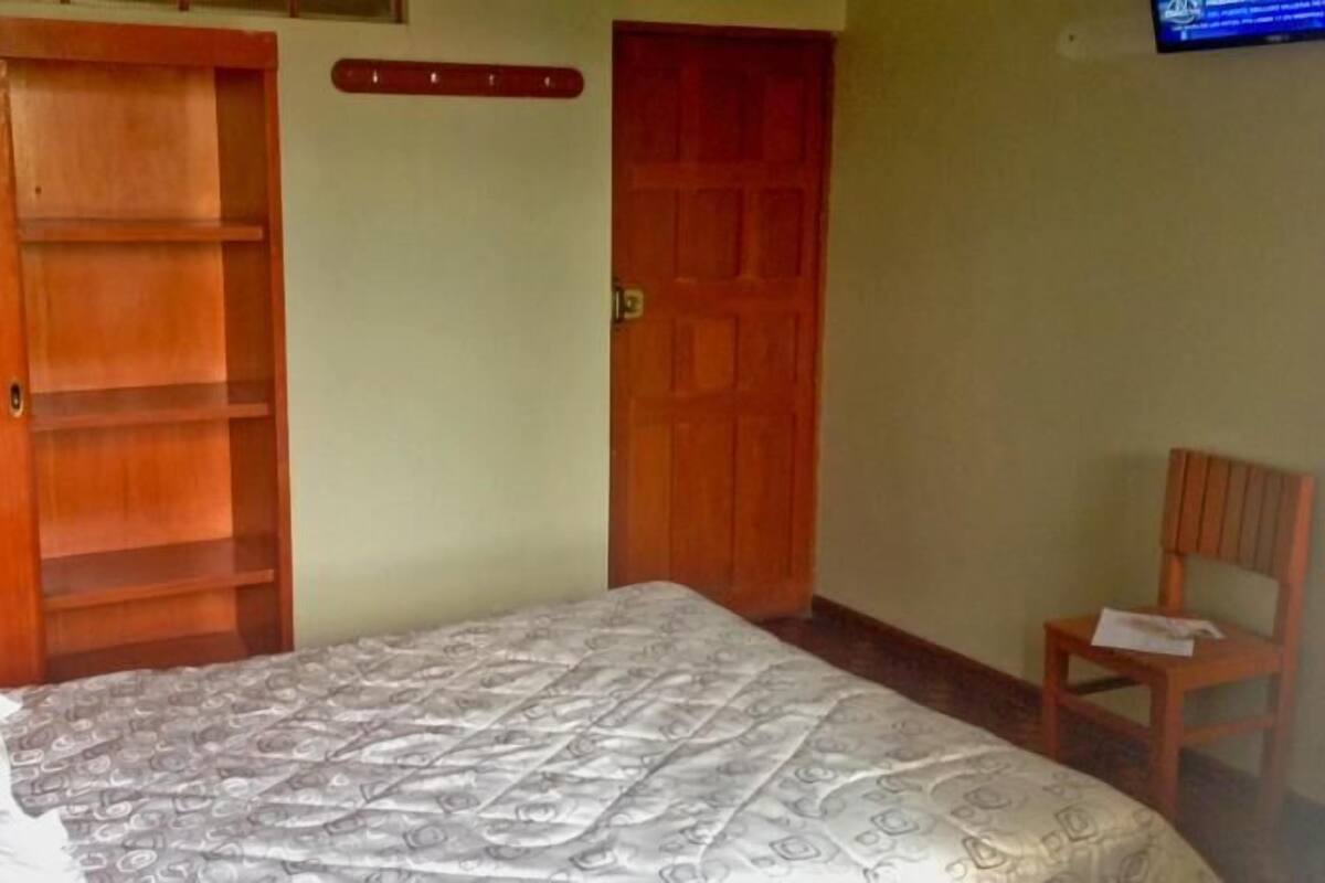 Quarto com cama, guarda-roupa de madeira, cadeira de madeira e televisão na parede.
