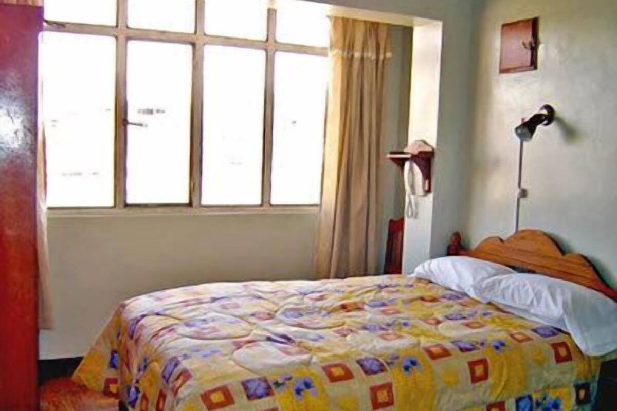 Quarto com cama, janela com cortinas, abajur e telefone fixado na parede.