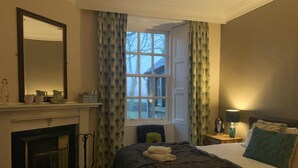 A bedroom with a large window, a fireplace, a bed with a dark blanket, and a bedside table with a lamp. - The Hopetoun Arms Hotel (Biggar)