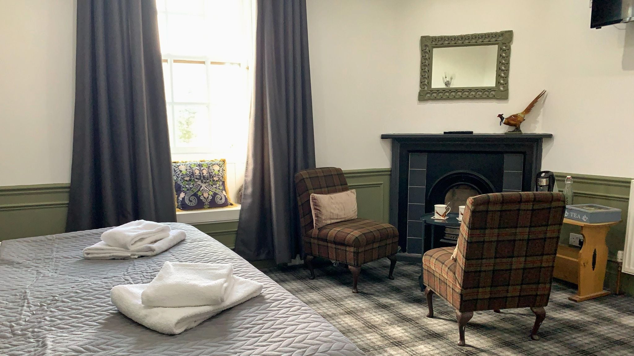 Habitación de hotel con cama, dos sillones a cuadros, chimenea y ventana con cortinas.