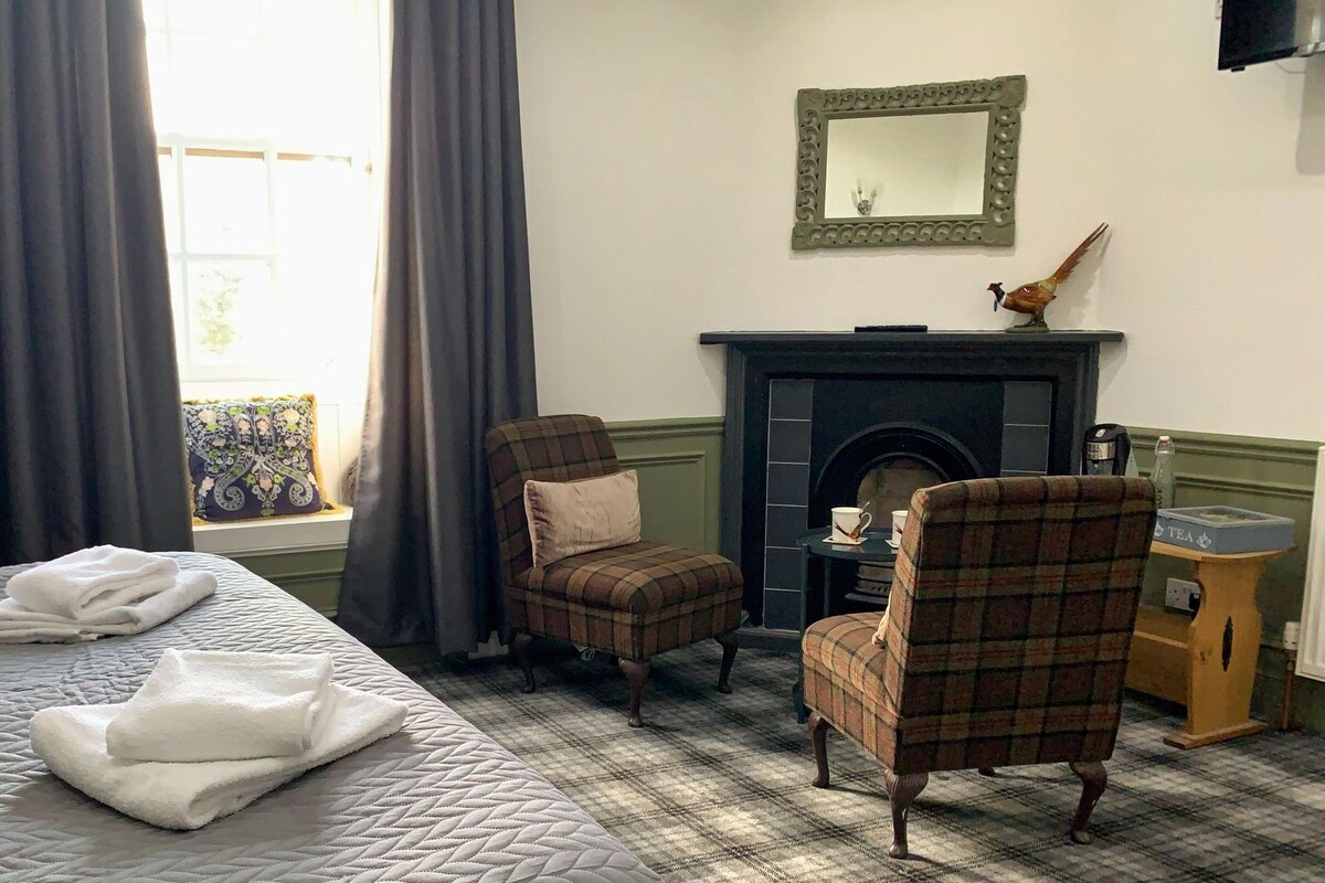 A hotel room with a bed, two plaid armchairs, a fireplace, and a window with curtains.