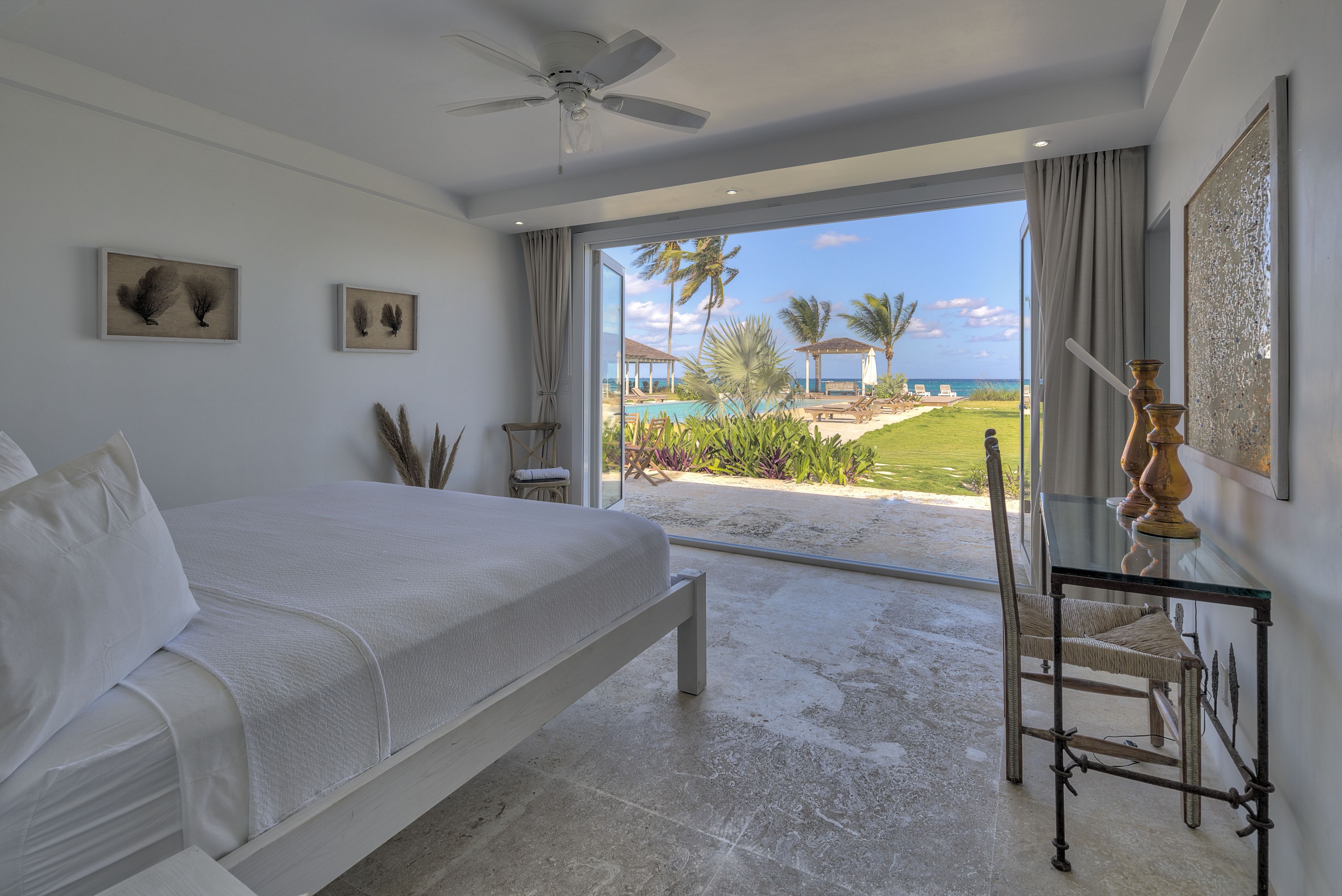 A bedroom with a bed, a ceiling fan, a glass table with a chair, and a view of a pool and palm trees.