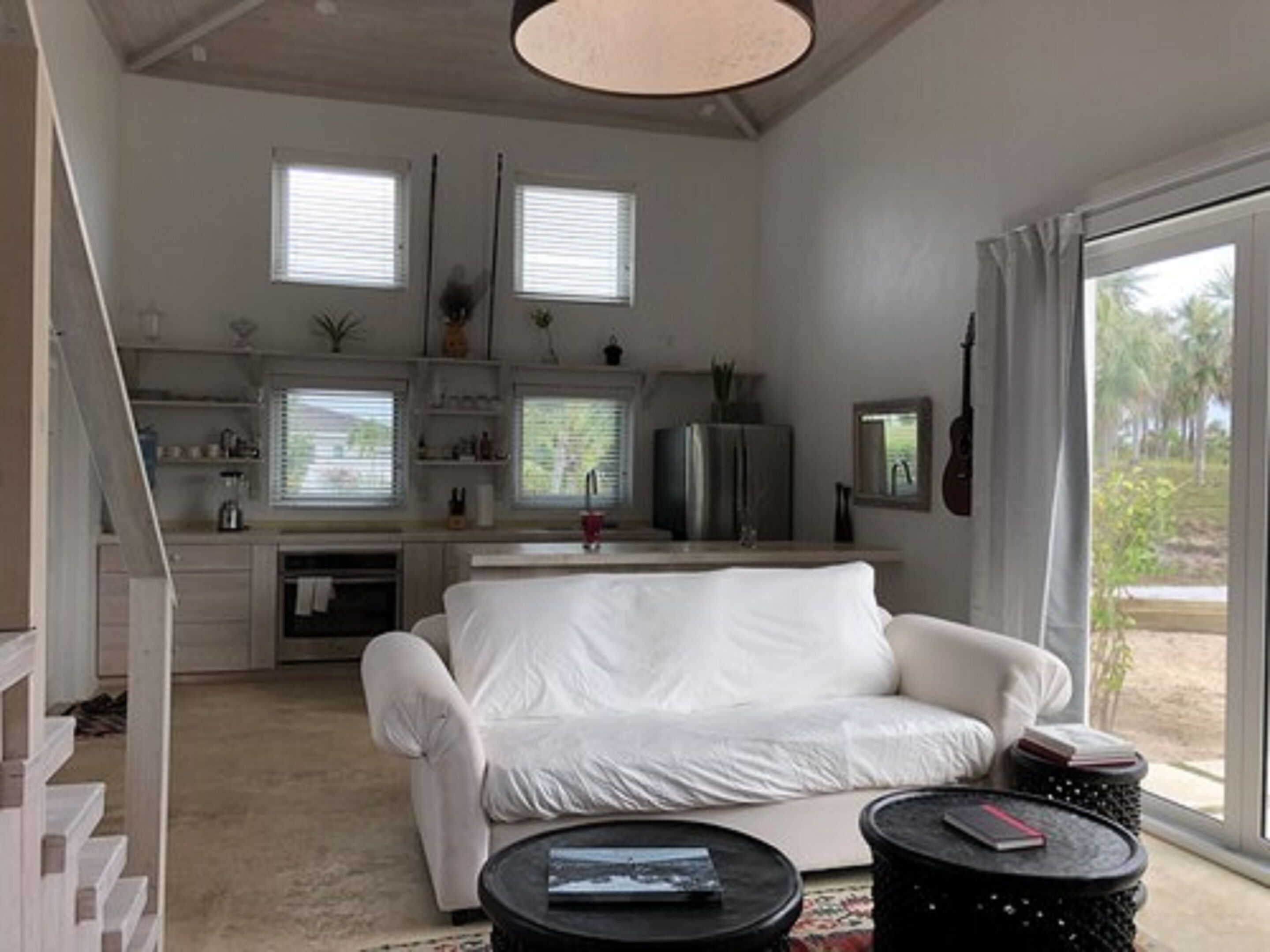 Een moderne woonkamer met een witte bank, een klein zwart bijzettafeltje en een keukenhoek met een koelkast en ramen.