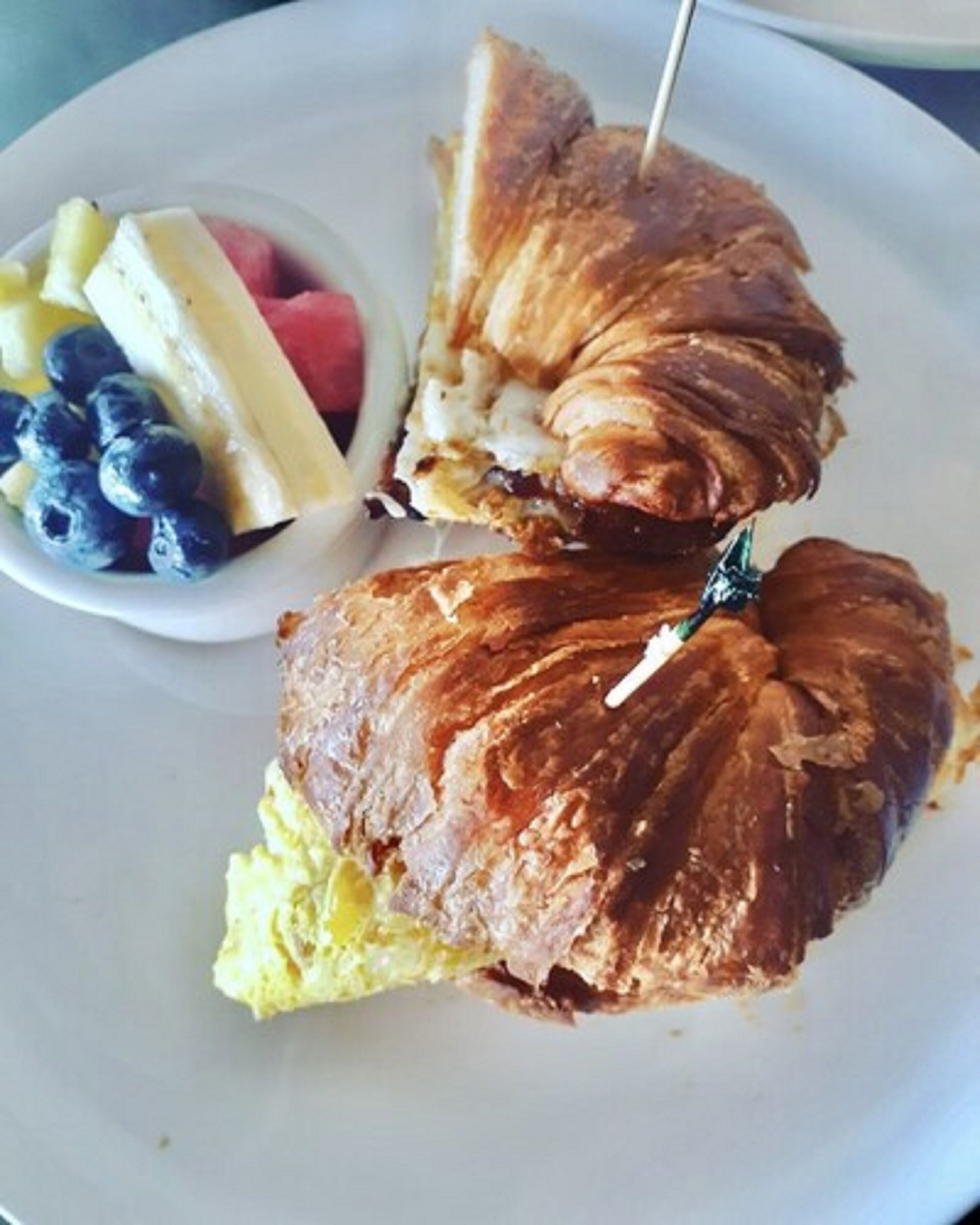 A plate with a croissant sandwich and a small bowl of fruit.