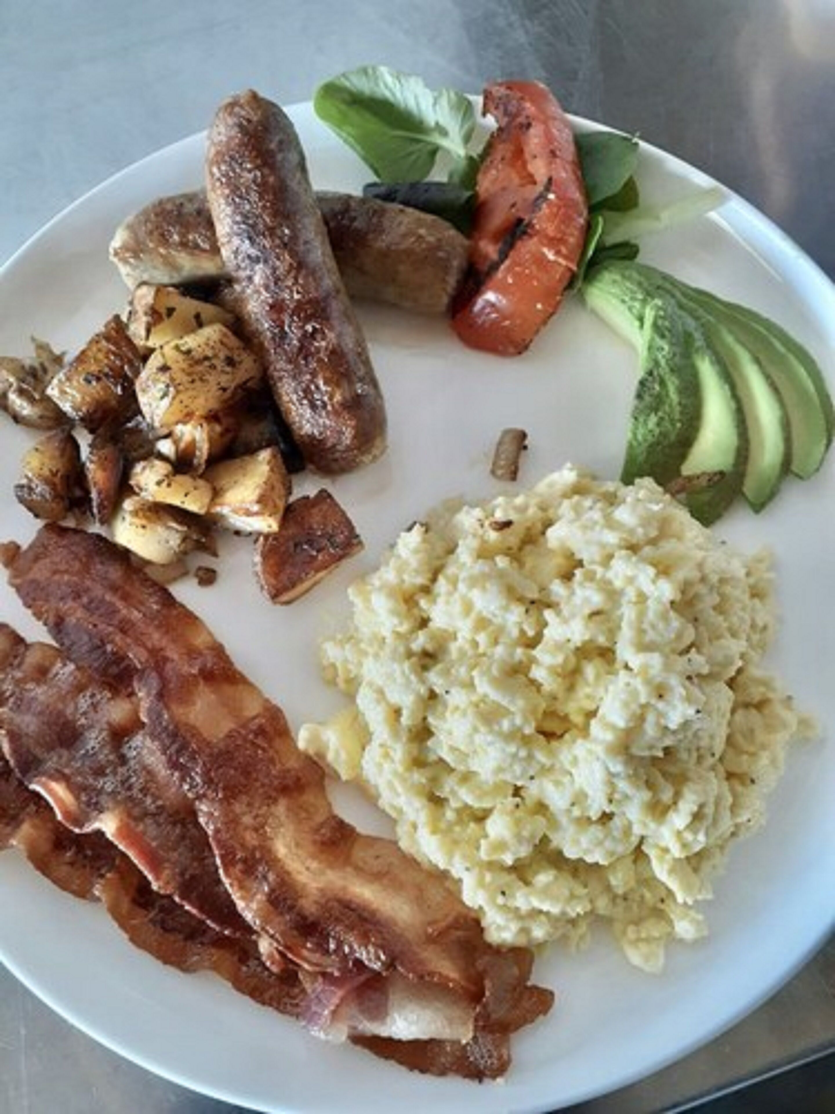 Une assiette contenant des œufs brouillés, du bacon, des saucisses, des pommes de terre rôties, des tomates grillées et des tranches d’avocat.