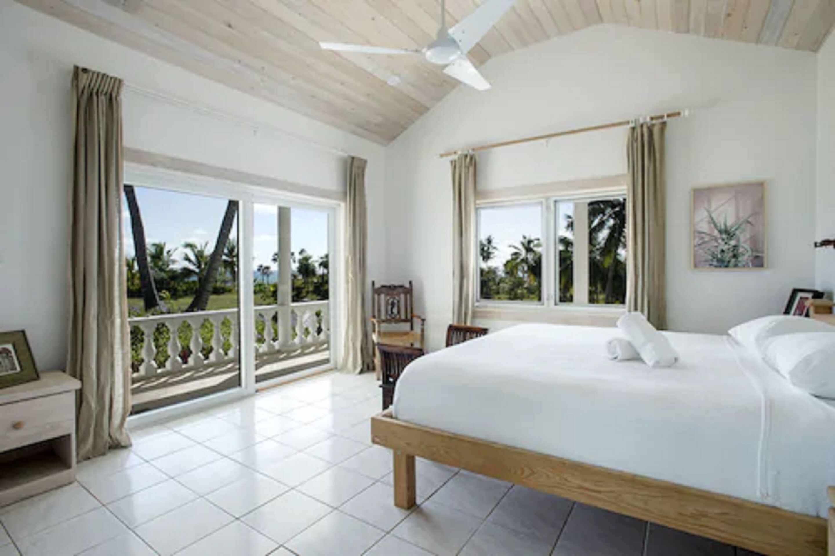 A bedroom with a large bed, a ceiling fan, and a view of palm trees through the windows.