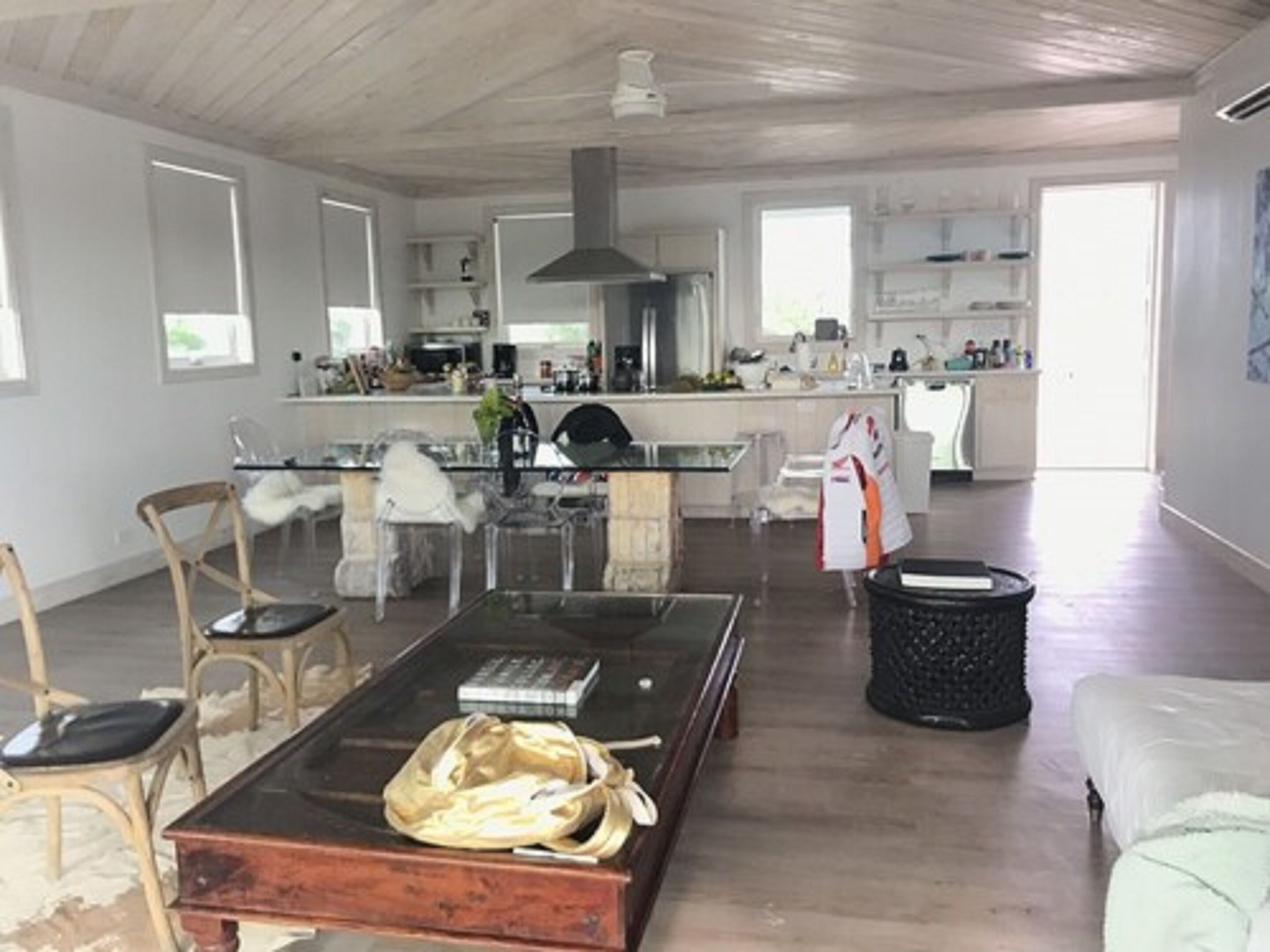 Un amplio salón con una mesa de centro de vidrio, una mesita auxiliar de madera y un sofá blanco. En el fondo, se ve una cocina con armarios blancos y una isla central.