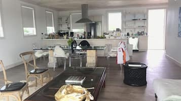 Un amplio salón con una mesa de centro de vidrio, una mesita auxiliar de madera y un sofá blanco. En el fondo, se ve una cocina con armarios blancos y una isla central.
