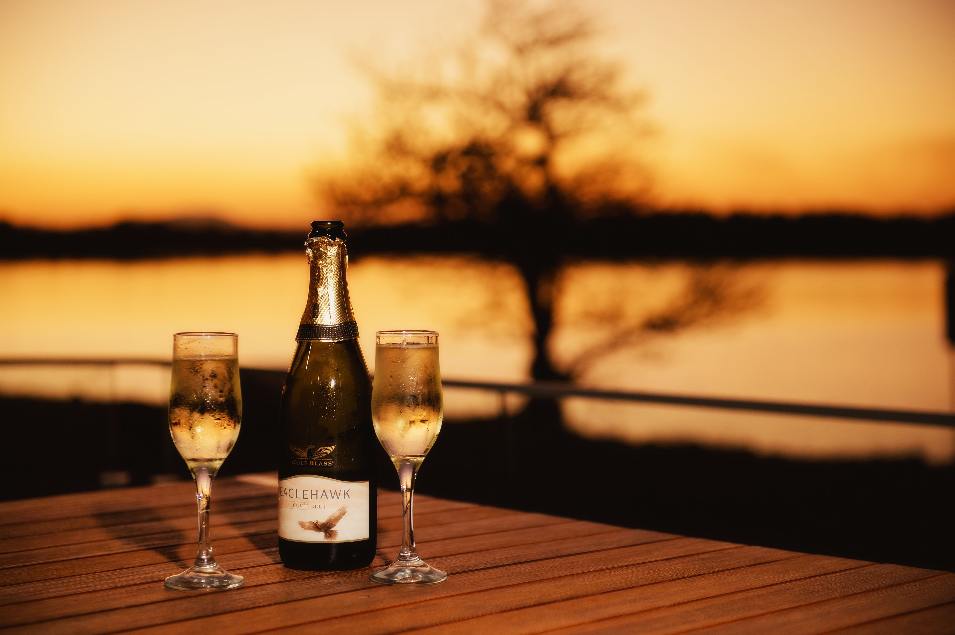 A bottle of Eaglehawk wine and two glasses on a wooden table with a sunset view in the background.