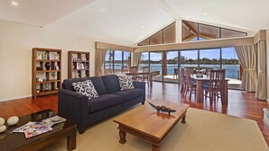 A spacious living room with a dark blue sofa, wooden coffee table, and bookshelves. A dining area with a table and chairs is visible through large windows overlooking a body of water. - Ana Mandara Luxury Retreat (Port Macquarie)