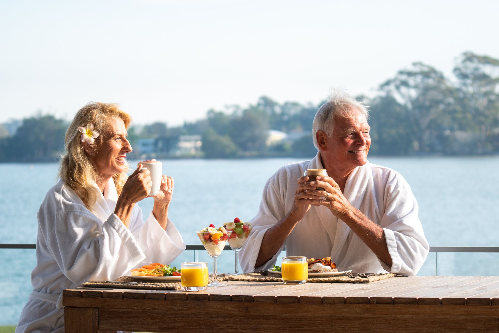 Two people sitting at a wooden table by a lake, enjoying breakfast with fruit and juice.