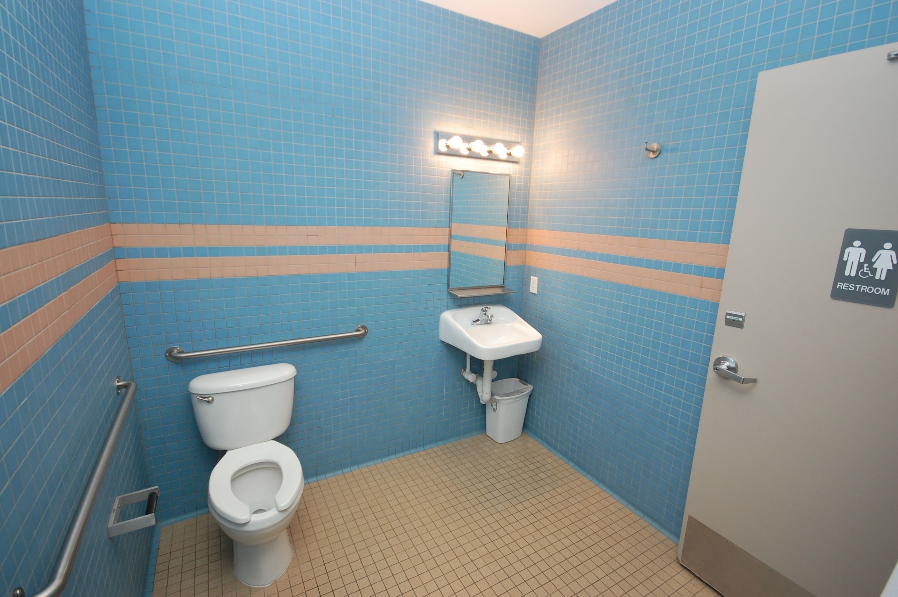 A restroom with blue tiled walls, a white sink, a mirror with lights, a handrail, and a sign indicating a handicap-accessible toilet.