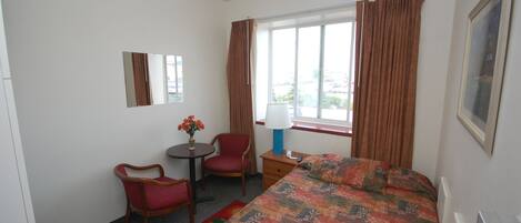 A hotel room with a bed, a desk with a chair, a small table with flowers, a lamp, and a window with curtains.