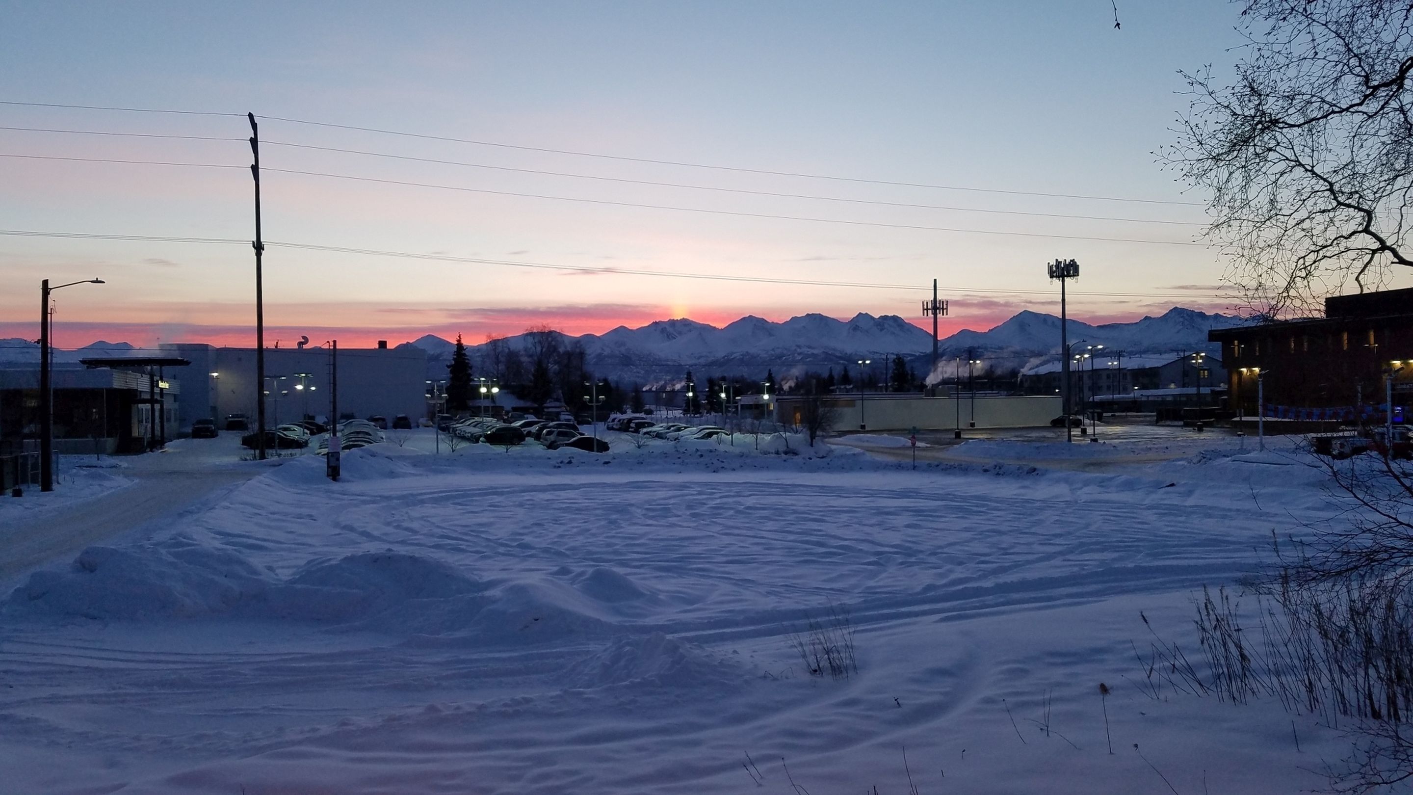 A snowy parking lot with cars and buildings in the background, surrounded by mountains.