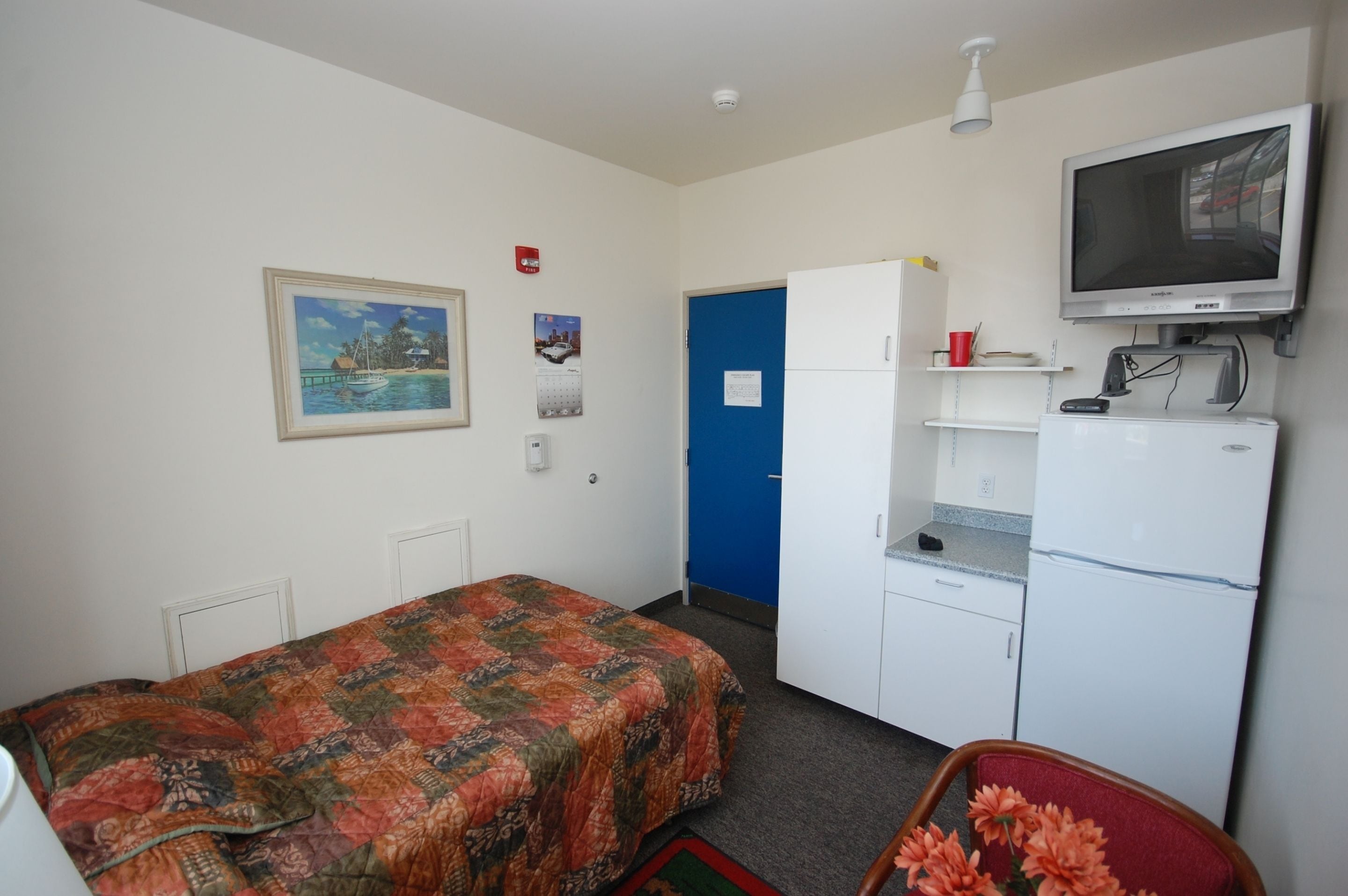 A small, compact room with a bed, a desk, a refrigerator, a television, and a blue door.