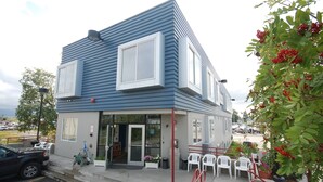 Un edificio de dos pisos con fachada azul, ventanas amplias y un pequeño espacio al aire libre con sillas blancas.