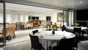 Uma ampla sala de jantar com várias mesas e cadeiras, cozinha ao fundo e grandes janelas que proporcionam iluminação natural.