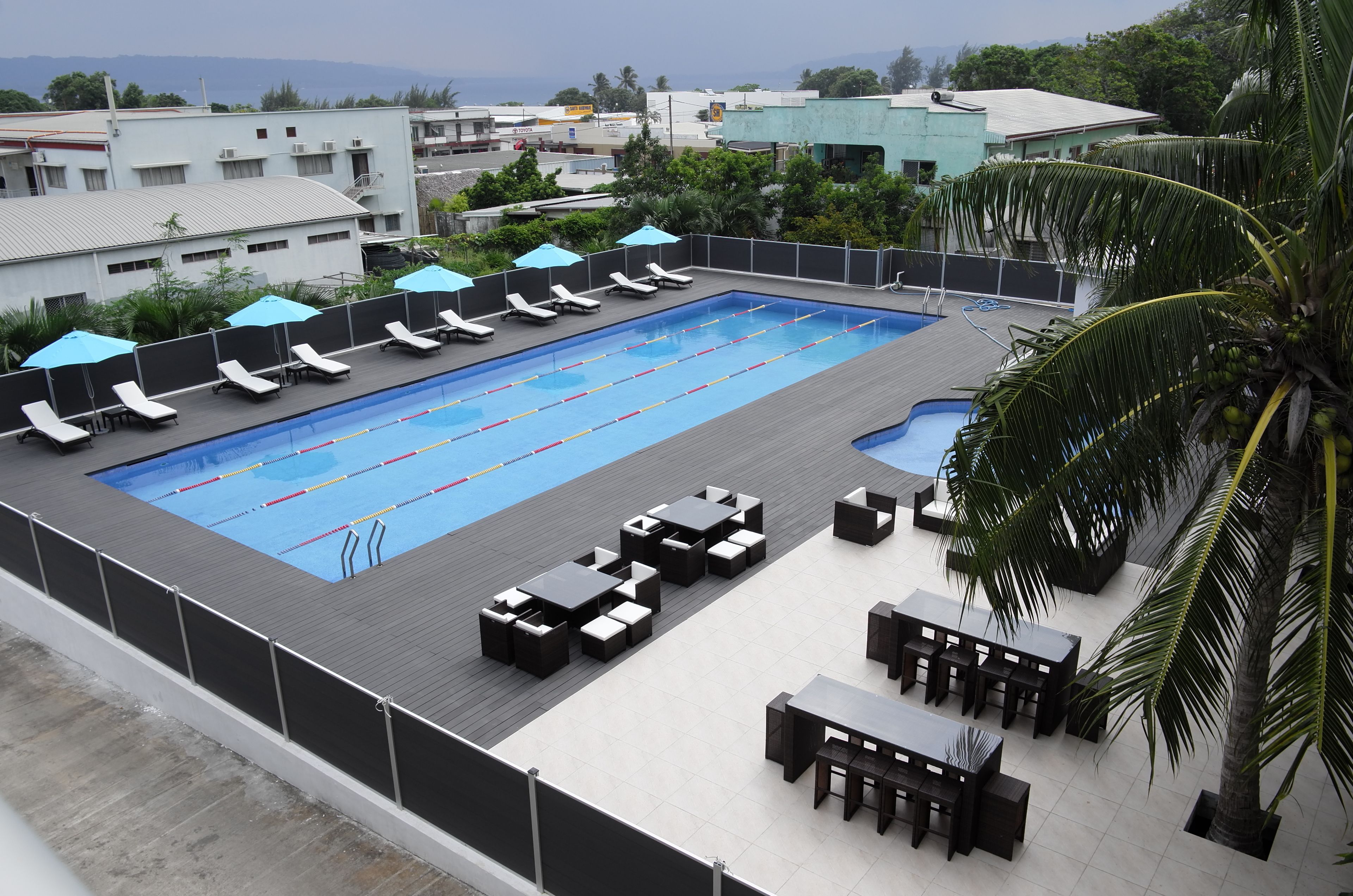 Une piscine sur le toit, agrémentée de chaises longues et de tables, et entourée de palmiers et d’immeubles.