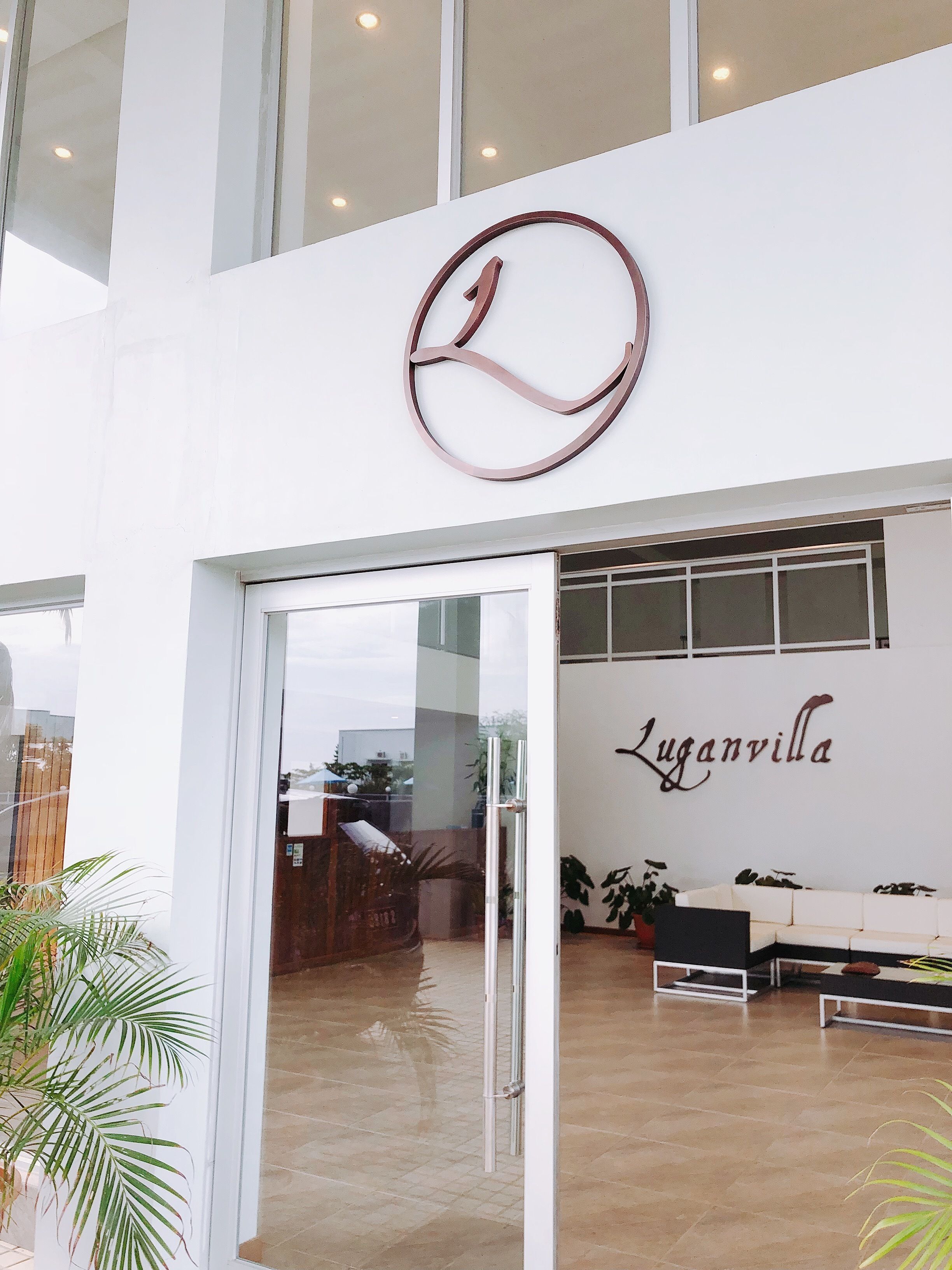 Una entrada moderna con puerta de vidrio, un letrero con logotipo circular y una pared con el nombre 'Luganvilla'.