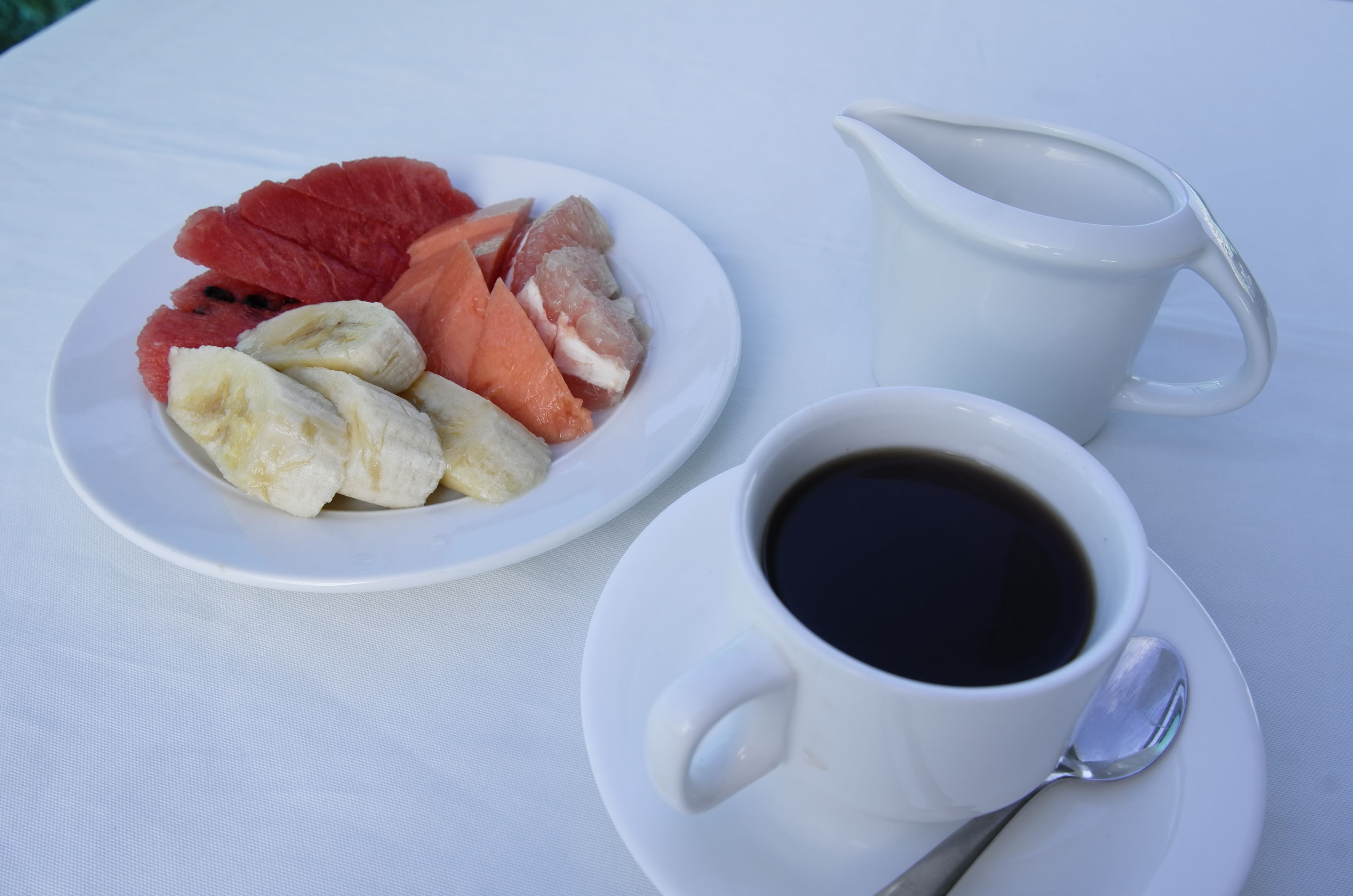 A white plate with sliced fruits and a cup of black coffee on a white tablecloth.