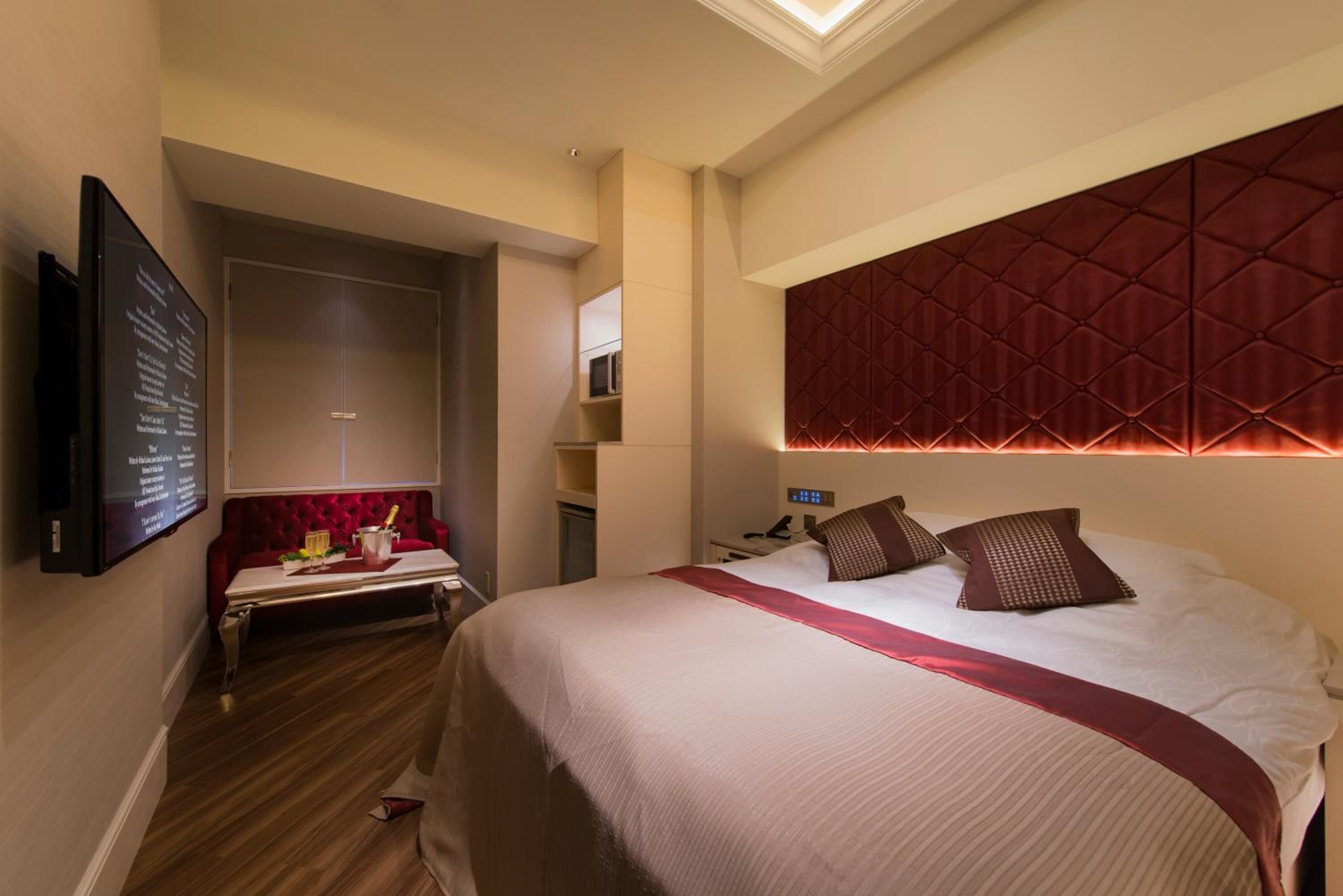 A hotel room with a large bed, a red upholstered bench, a flat-screen TV mounted on the wall, and a built-in headboard with a diamond-patterned design.