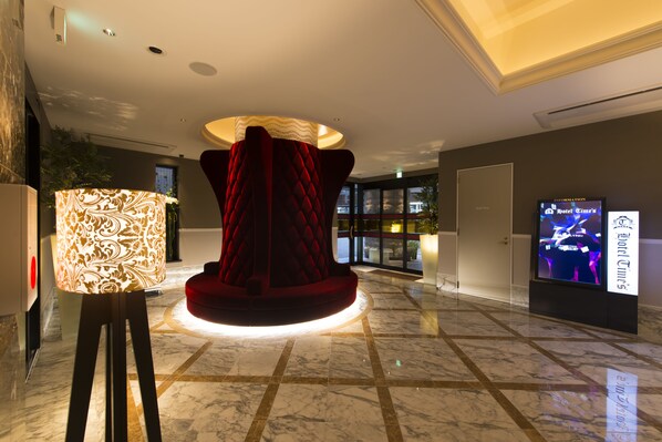 A lobby with a central red velvet chair, a patterned floor lamp, and a digital screen displaying hotel information.