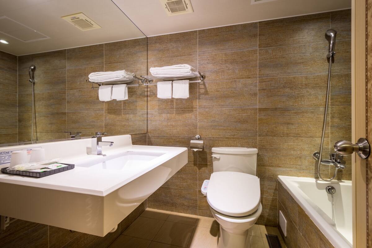 A modern bathroom with a large white sink, a bathtub, and a toilet. There are towels stacked on a rack above the sink.