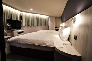A modern hotel room with a large bed, a flat-screen TV, a bedside table, and a window with curtains.