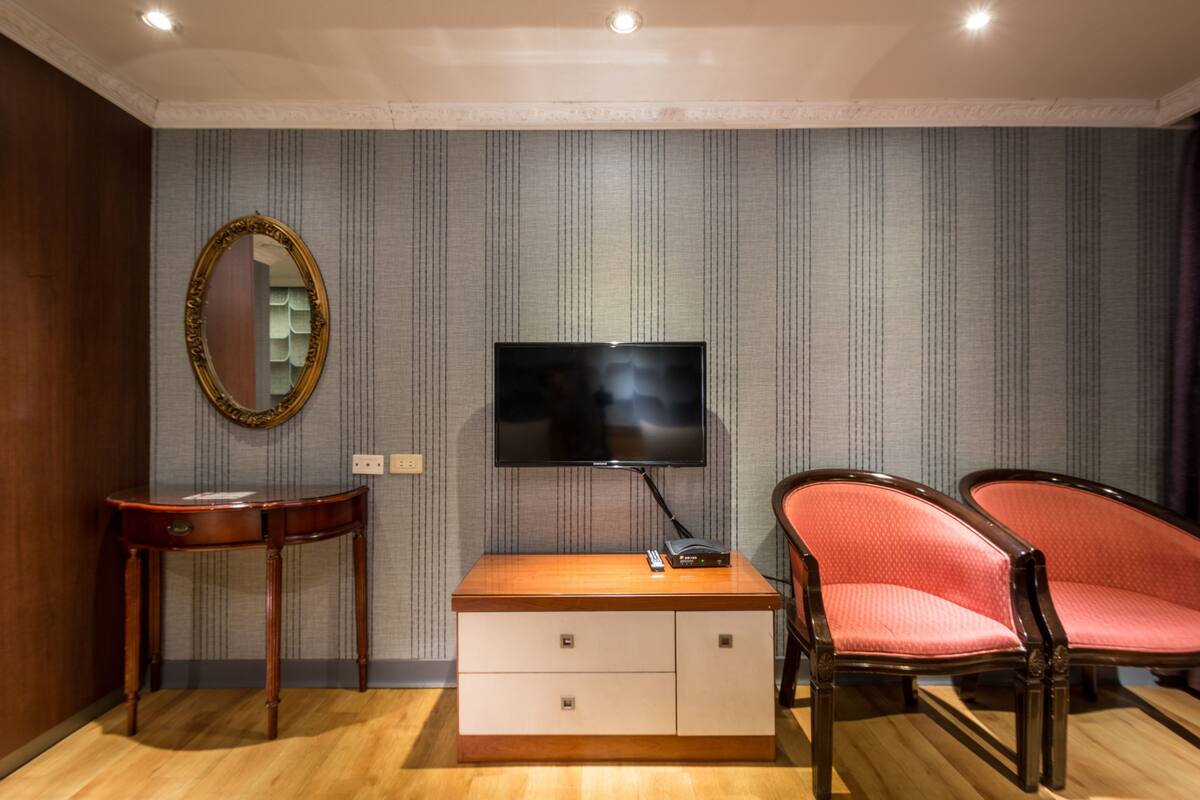A hotel room with a wooden dresser, a television, a mirror, and two upholstered chairs.