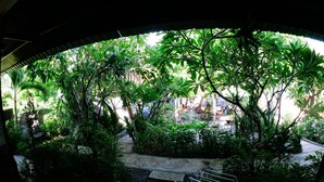 A view through a curved window of a lush garden with a paved area and a metal bench. - Sea Hua Ha Ha - Hostel (Tulamben)