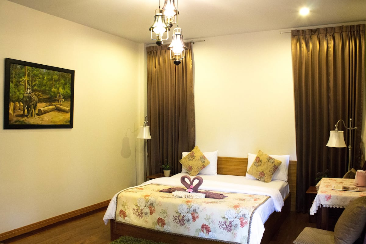 A hotel room with a large bed, two bedside tables, a nightstand, a desk with a chair, a potted plant, and a painting on the wall.