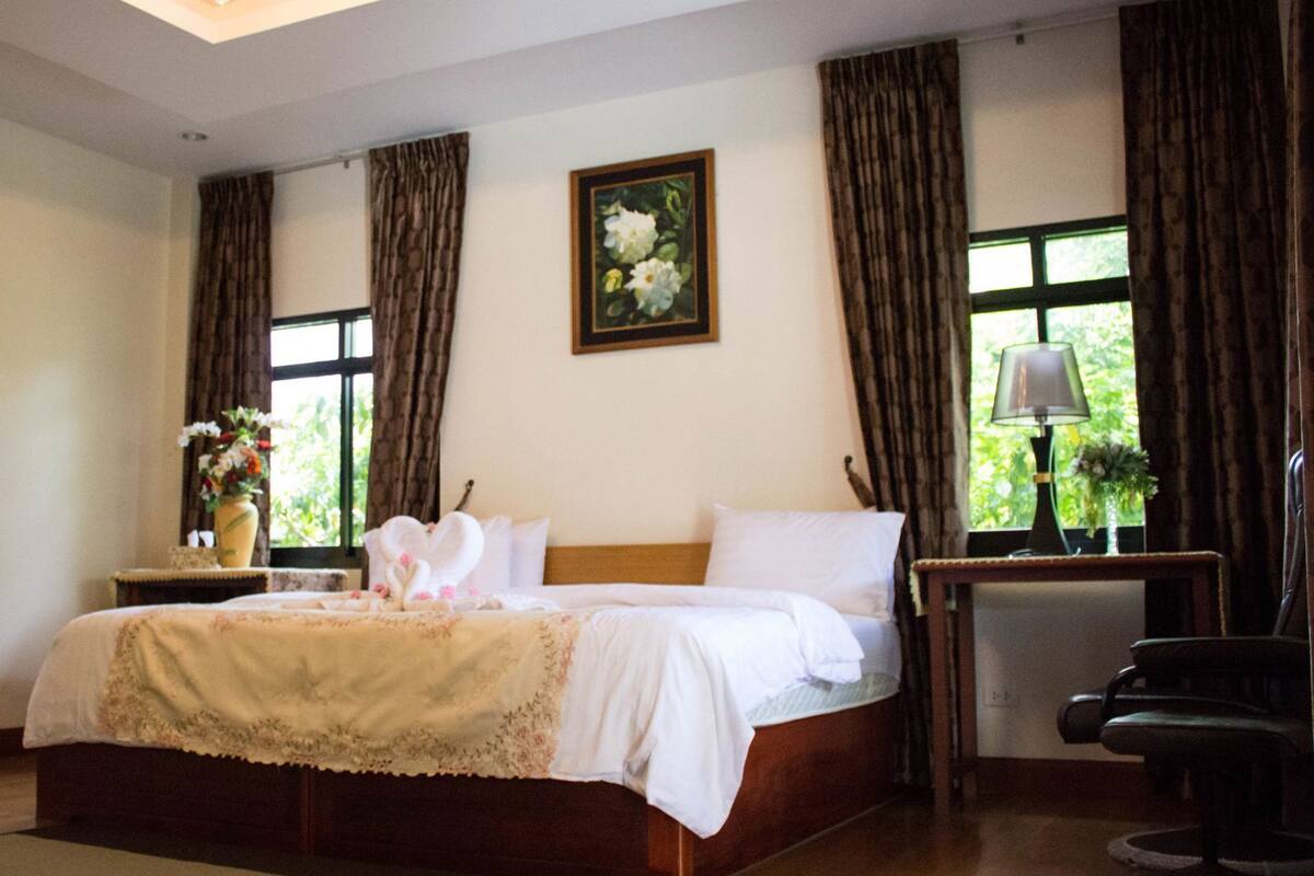 A bedroom with a bed, a nightstand, a chair, a chandelier, a window with curtains, and a painting on the wall.