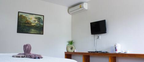 A hotel room with a bed, a painting, a television, a small plant, and a wooden side table with a kettle and cups.