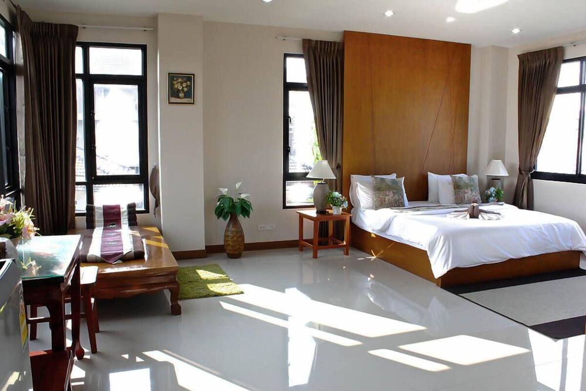 A hotel room with a large bed, a desk, a chair, a small table, a plant, and a window with curtains.