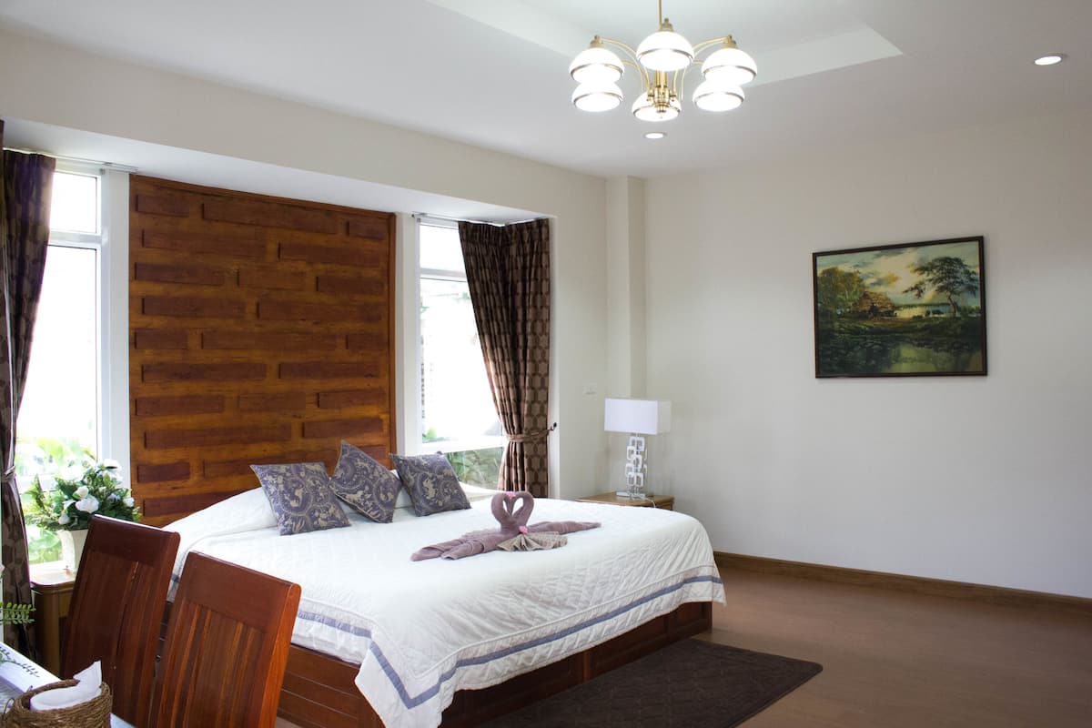 A bedroom with a large bed, wooden headboard, a nightstand with a lamp, a painting on the wall, and a window with curtains.