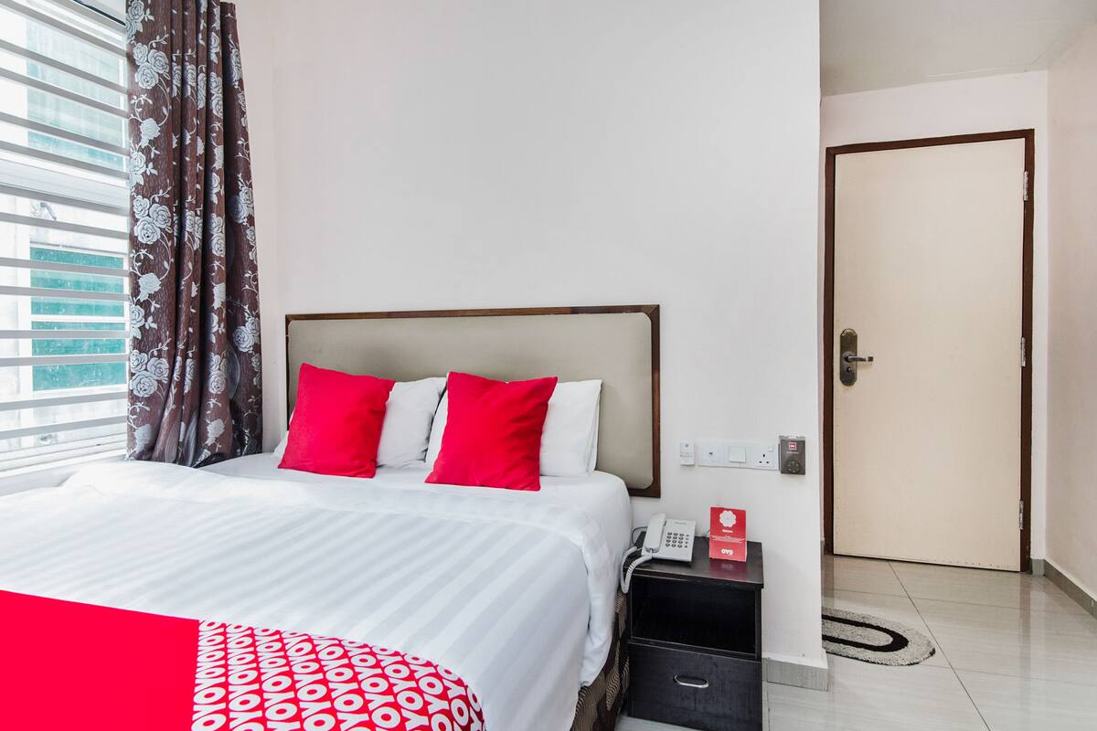 A hotel room with a bed, red and white pillows, a bedside table with a phone and a red box, and a door.