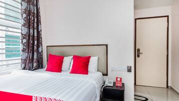 A hotel room with a bed, red and white pillows, a bedside table with a phone and a red box, and a door.