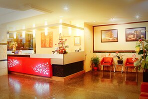A hotel reception area with a red reception desk, a large mirror, framed pictures on the wall, and two red chairs with potted plants.