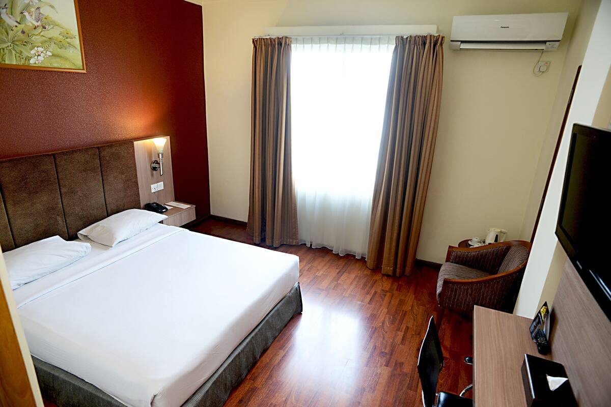 A hotel room with a large bed, a desk, a chair, an air conditioner, and a window with curtains.