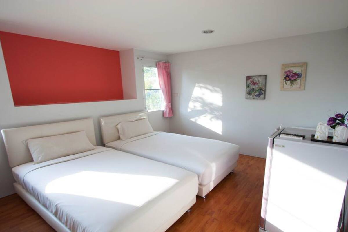 Una habitación con dos camas, una pared de acento roja, una ventana con cortinas rosadas y un refrigerador con flores.