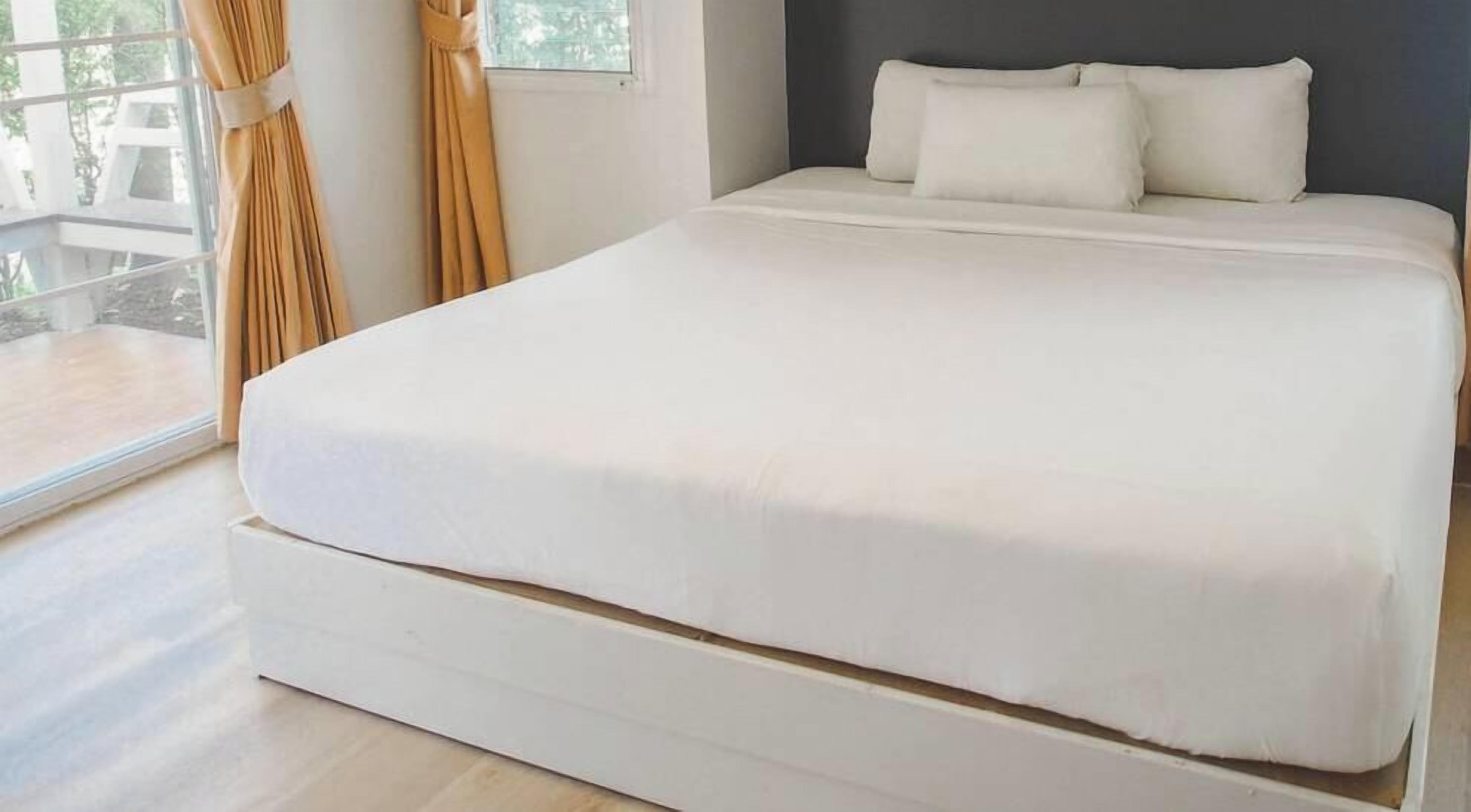 A large bed with a white mattress and pillows, a window with curtains, and a balcony outside.