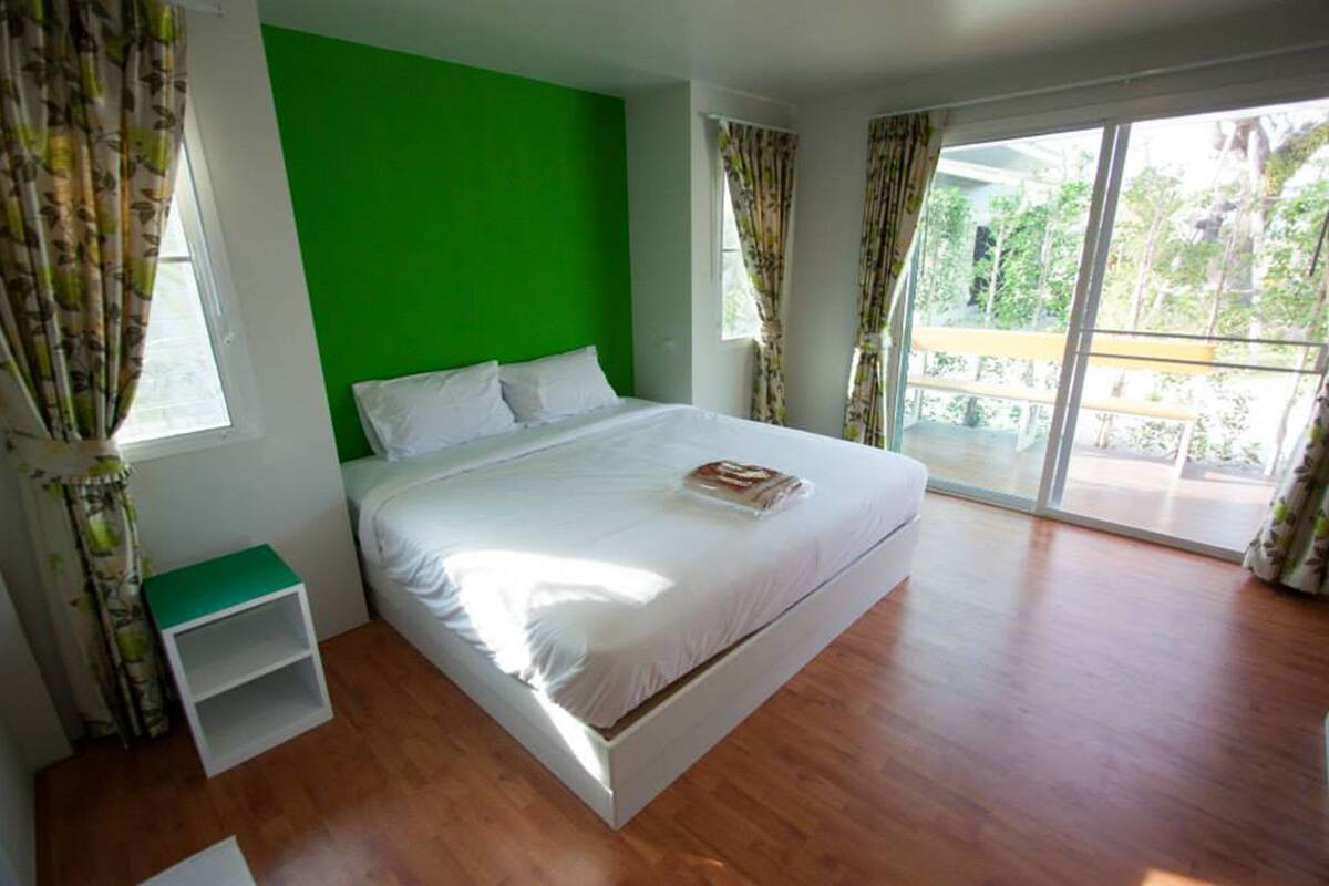 Quarto moderno com parede verde, uma cama grande, uma mesa de cabeceira branca e uma varanda com vista para o verde.