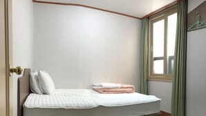Una cama individual con cabecera de madera, colcha blanca y dos almohadas. Una cortina verde cubre parcialmente una ventana a la derecha.