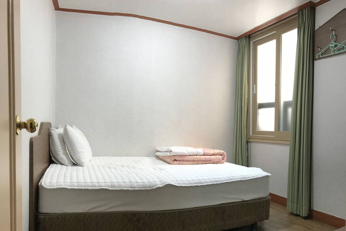 Una cama individual con cabecera de madera, colcha blanca y dos almohadas. Una cortina verde cubre parcialmente una ventana a la derecha.